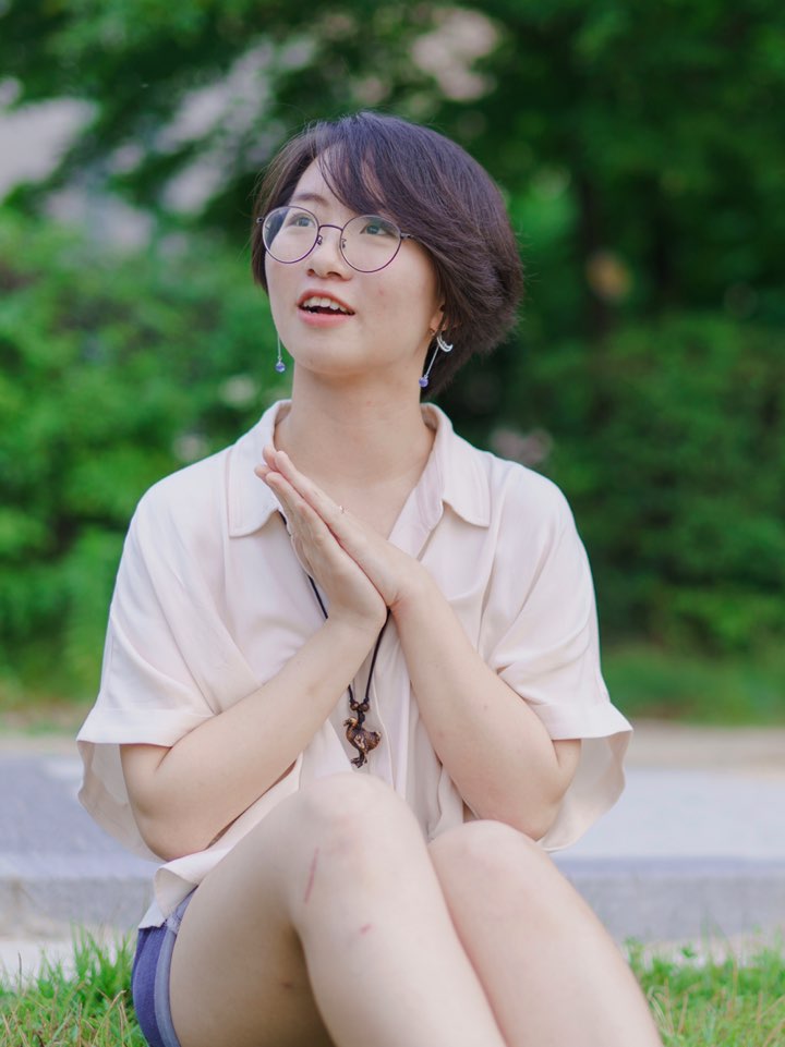 Picture of me with short hair and round glasses, sitting in a meadow with a Dodo bird necklace.